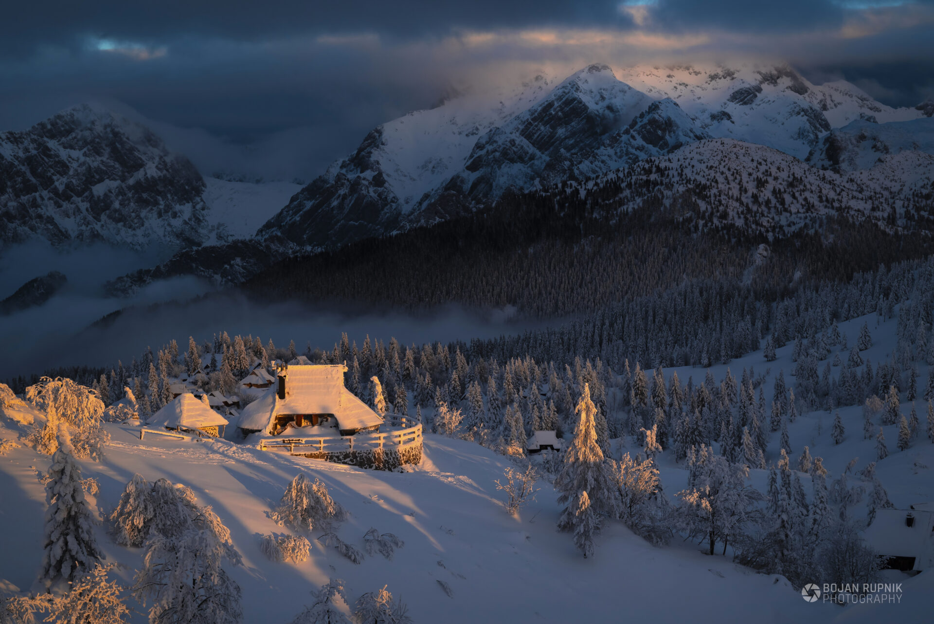 Koča na zasneženi Veliki Planini, obsijana z jutranjim soncem soncem in Kamniškimi Alpami v ozadju.